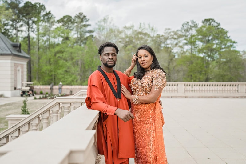 A couple stands together outdoors at Oxbow Estate, dressed in elegant traditional attire. The man wears a red and black outfit, and the woman wears an ornate orange dress with lace details. They