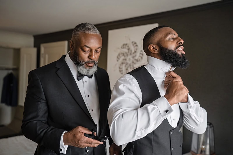 Two men in formal attire prepare in a room; one adjusts the other's bow tie while holding a phone, both wearing black suits with suit buttons done and white shirts. They exhibit focused and joyful expressions