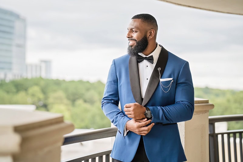 A man in a stylish one-button blue suit and bow tie stands on a balcony. He has a full beard, smiles gently, and adjusts his cufflinks. Trees and a building are visible in the