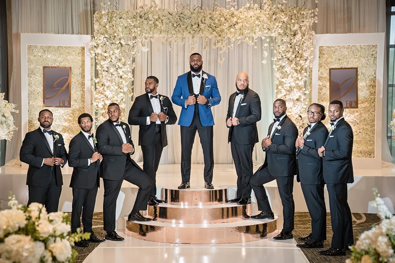 Eight men in dark tailored suits adhering to the classic three-button rule posing confidently in front of an elegantly designed wedding stage framed by floral arrangements, with mirrored accents and warm lighting.