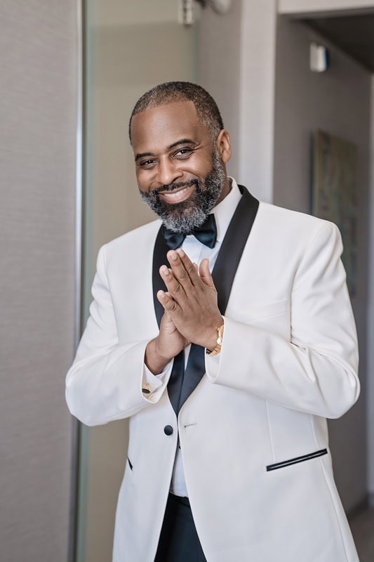 A middle-aged man in a stylish white tuxedo with black lapels follows the suit button rule of one button and sports a black bow tie. He smiles warmly, has a short beard, and