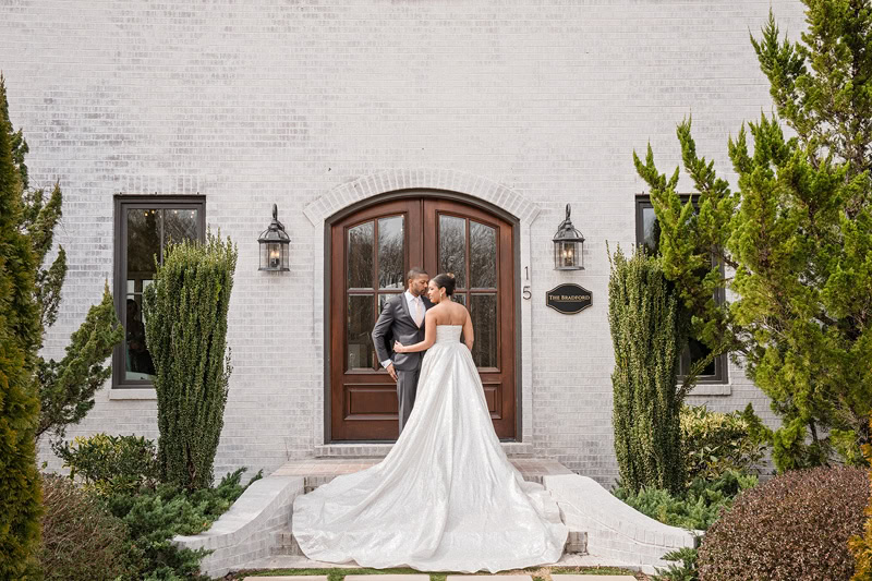 A bride and groom share a kiss on the steps of The Bradford wedding venue, flanked by tall green shrubs and exterior lanterns. The bride's long, flowing dress trails elegantly down the