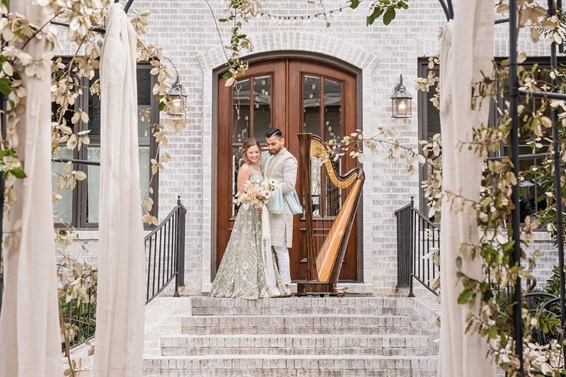A newlywed couple poses on stone steps in front of an arched wooden door at The Bradford wedding venue, flanked by white flowers and a harp. The bride holds a bouquet; both are