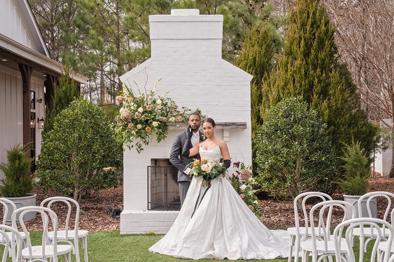 A bride and groom stand in front of a white brick fireplace adorned with flowers, surrounded by empty white chairs and green shrubbery, at The Bradford wedding venue.
