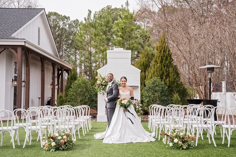 A newlywed couple smiling outdoors at The Bradford wedding venue, with the bride in a long white gown and the groom in a classic black suit. Chairs line the aisle, flanked by floral arrangements,