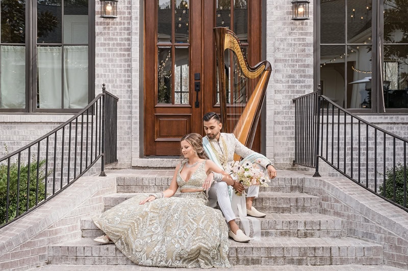 A bride and groom sit together on stone steps outside The Bradford wedding venue with large windows and doors. The bride is in an ornate gown, and the groom wears a white suit. A large har