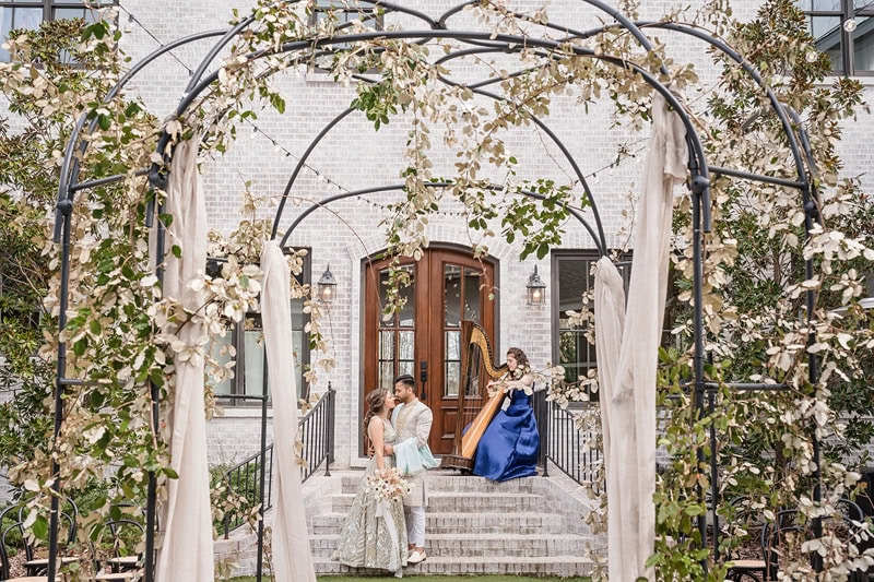 A wedding scene at The Bradford wedding venue, with a couple seated on steps under an arched trellis adorned with white flowers. The bride in a long white gown and the groom in a suit