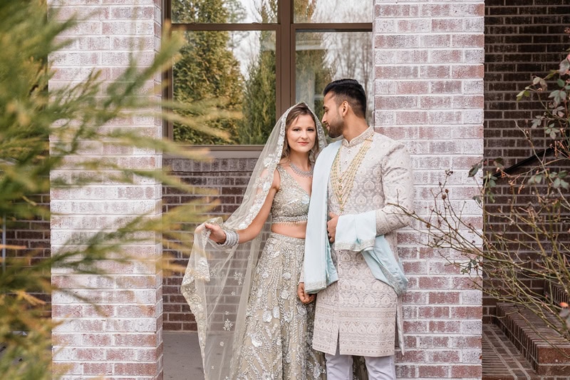 A bride and groom in traditional South Asian wedding attire stand affectionately together between brick columns at The Bradford wedding venue, surrounded by greenery. The bride wears a detailed white and silver lehenga,