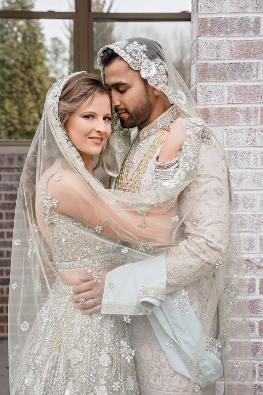 A bridal couple embracing under a translucent veil at The Bradford wedding venue, with the bride in a beaded gown and the groom in a detailed cream sherwani, against a brick wall background.