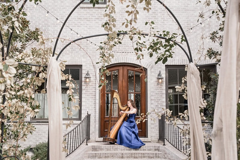A woman in a blue gown plays a harp under a vine-covered archway leading to The Bradford wedding venue with elegant wooden doors, surrounded by draping greenery and wispy curtains, creating a