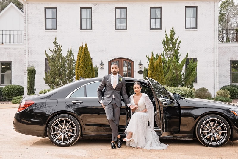 A bride in a white gown and groom in a gray suit standing beside a black Mercedes-Benz at The Bradford wedding venue with pointed trees lining the doorway.