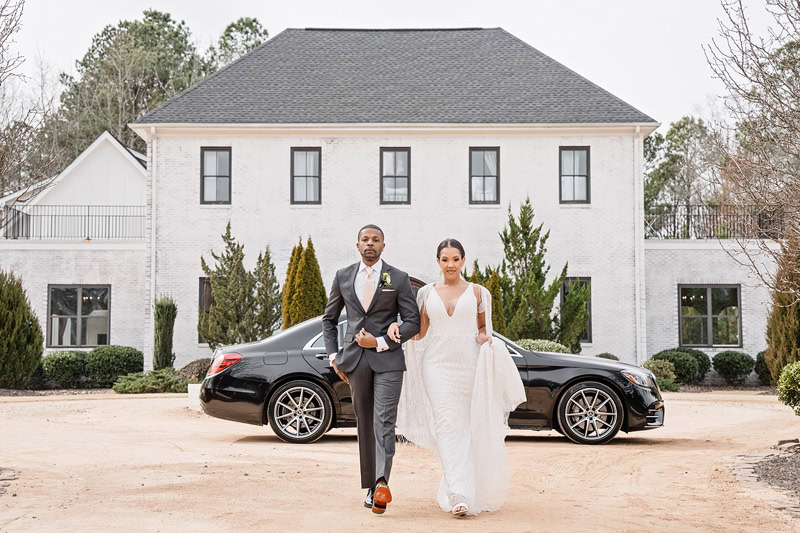 A bride and groom walk hand-in-hand in front of The Bradford wedding venue, a large, white two-story house, with a black luxury car parked beside them. They are elegantly dressed, the