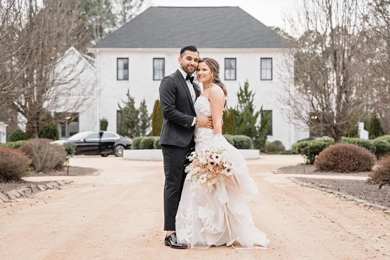 A newlywed couple stands on a gravel path, embracing in front of the large, white Bradford wedding venue. The man wears a black suit and the woman is in an elegant white wedding dress with a
