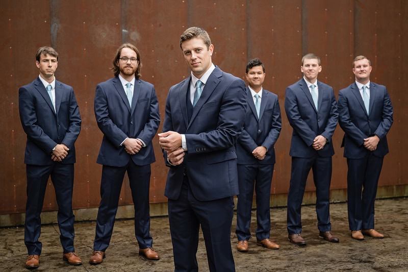 A group of seven men in matching blue three-button suits and brown shoes, standing confidently before a rustic brown background. The man in front is centrally positioned, staring intently at the camera, with the