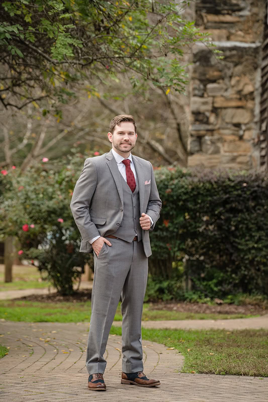 A man in a tailored two-button light gray suit and red tie stands confidently on a paved pathway in a park. He has short brown hair and a beard, and holds his jacket with one hand,