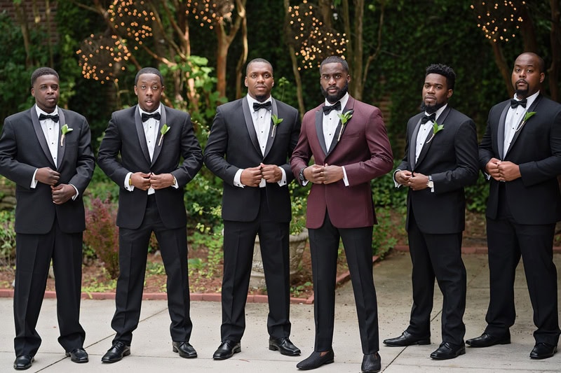 Five men in formal attire pose outdoors. Four wear black tuxedos with white shirts and bow ties, adhering to the traditional suit button rule, while the central figure stands out in a mar